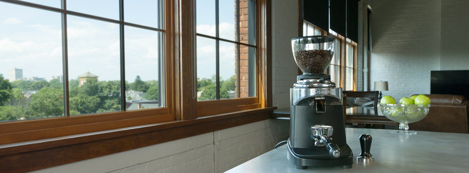 A commercial coffee and espresso grinde at the edge of a white countertop beside a woodgrain framed window.