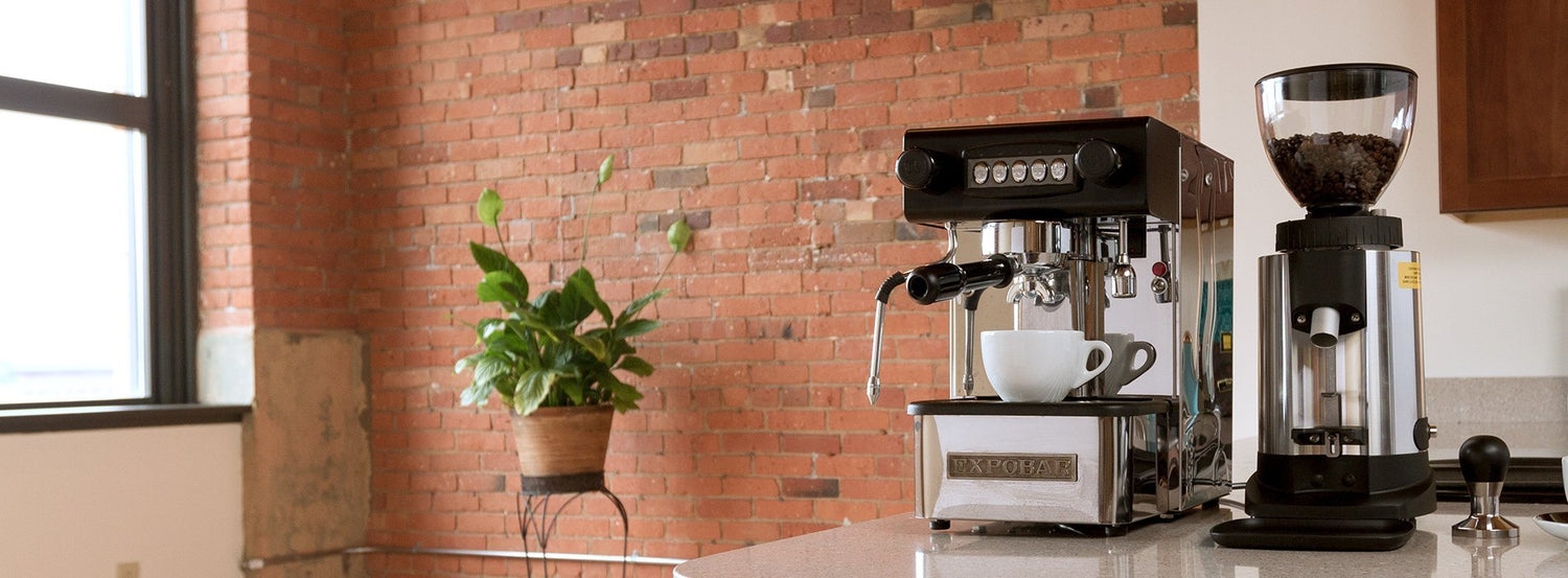 A rebuilt espresso machine next to an espresso grinder on a kitchen counter in front of a brick wall.