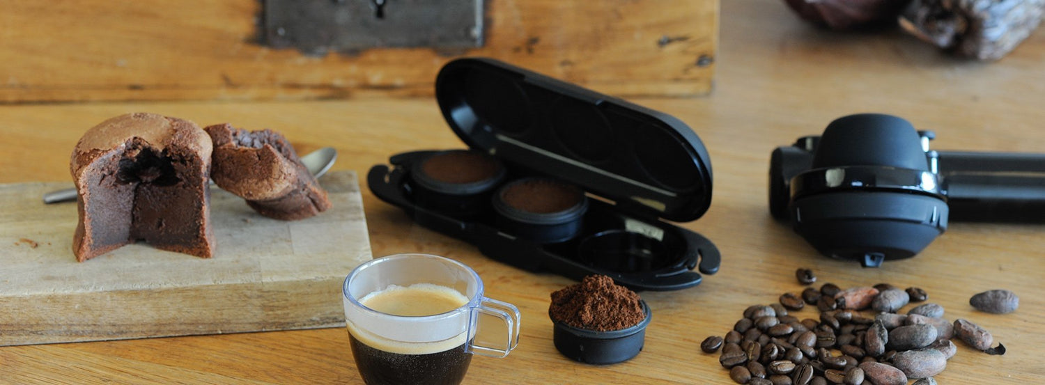 A Handpresso manual espresso machine next to coffee beans, ground coffee, and a cup of espresso.