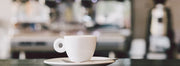 An espresso cup and saucer set in front of an out of focus commercial espresso machine and grinder