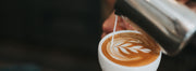 Milk pouring from a frothing pitcher into a cup of espresso to form latte art of a frond.