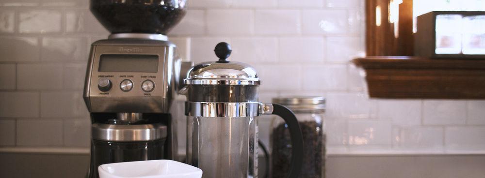 A french press in front of a coffee grinder and container full of coffee beans.