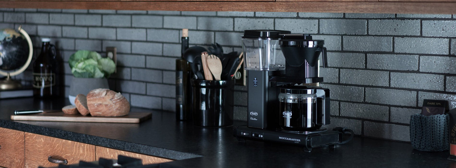 A Technivorm coffee maker on a black counter in front of a gray brick backsplash.