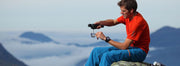 A man in a red-orange shirt sitting on a rocky ledge brewing espresso with a Handpresso.