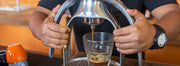 A ROK Espresso Maker brewing espresso into a clear espresso cup in front of a bright orange background.