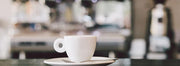 A white coffee cup and saucer set in front of an out of focus espresso machine and coffee grinder.