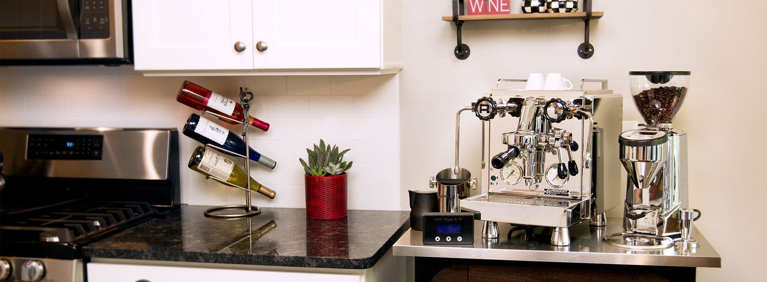 A Rocket Espresso machine and coffee grinder on a small island next to a black kitchen counter and white cabinets.
