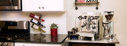 A Rocket Espresso machine and coffee grinder on a small island next to a black kitchen counter and white cabinets.