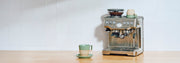 Breville Barista Touch on a wooden counter next to a green and brown coffee cup.