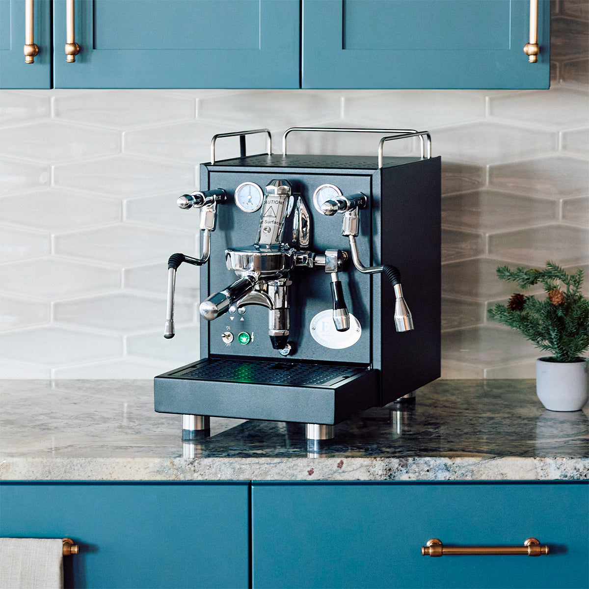 Black Mechanika Max Espresso machine on a kitchen counter with blue cabinets and a plant in the background.
