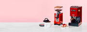 Red Gaggia Classic E24 next to a Red Eureka Silenzio 55 in front of a pink background and on a white counter.
