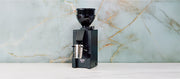 Close-up of a black Eureka Mignon Zero 55s on a white counter in front of a blue slate wall. 