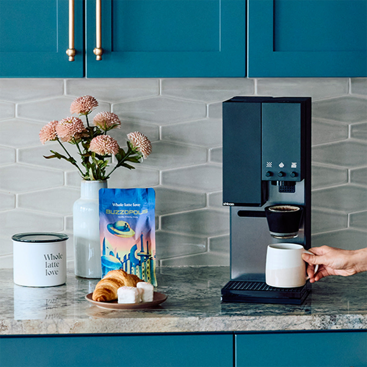 Close-up of the xBloom Studio in Black on a kitchen counter with blue cupboards next to  Whole Latte Love Buzzopolis, coffee canister, pastry and flowers.