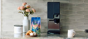Close-up of the xBloom Studio in Black on a kitchen counter with blue cupboards next to  Whole Latte Love Buzzopolis, coffee canister, pastry and flowers.
