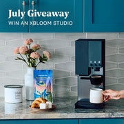 xBloom Studio in Black on a kitchen counter with blue cupboards next to  Whole Latte Love Buzzopolis, coffee canister, pastry and flowers. Promotional text overlayed that reads "July Giveaway - Win an XBloom Studio".