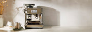 A Breville Barista Series espresso machine in brushed stainless steel, shown on a countertop with a milk frothing pitcher, stacked cups, and a green cloth against a tiled wall.