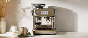 Close-up of a Breville Barista Series espresso machine in brushed stainless steel, shown on a countertop with a milk frothing pitcher, stacked cups, and a green cloth against a tiled wall.