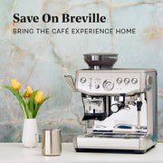 Close-up of the Breville Barista Express Impress next to yellow tulips on a white counter in front of a blue slate wall. Promotional text overlayed that reads "Save On Breville - Bring the cafe experience home".