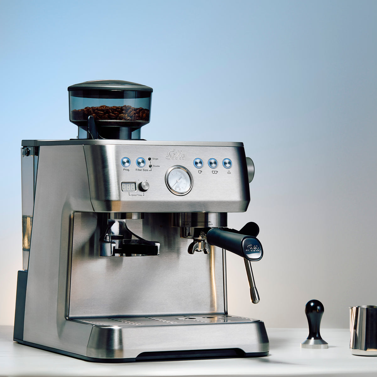 Close-up of a Solis Grind & Infuse Perfetta in Stainless Steel on a white counter top next to a tamper and milk frothing pitcher.