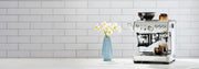 Breville Barista Express Impress next to flowers on a white counter in front of white a tiled wall.