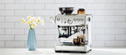 Close-up of the Breville Barista Express Impress next to flowers on a white counter in front of white a tiled wall.
