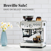 Breville Barista Express Impress next to flowers on a white counter in front of white a tiled wall. Promotional text overlayed that reads "Breville Sale! Save on select machines."