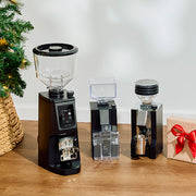 Three coffee grinders on a wooden surface with a Christmas tree and gift box in the background.